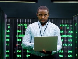 Male system administrator of big data center typing on laptop computer while working in server room. Programming digital operation. Man engineer working online in database center. Telecommunication.