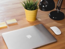 Closed MacBook on a desk with two sticky note pads, an Apple mouse, a desk lamp, and a small potted plan arranged around it.